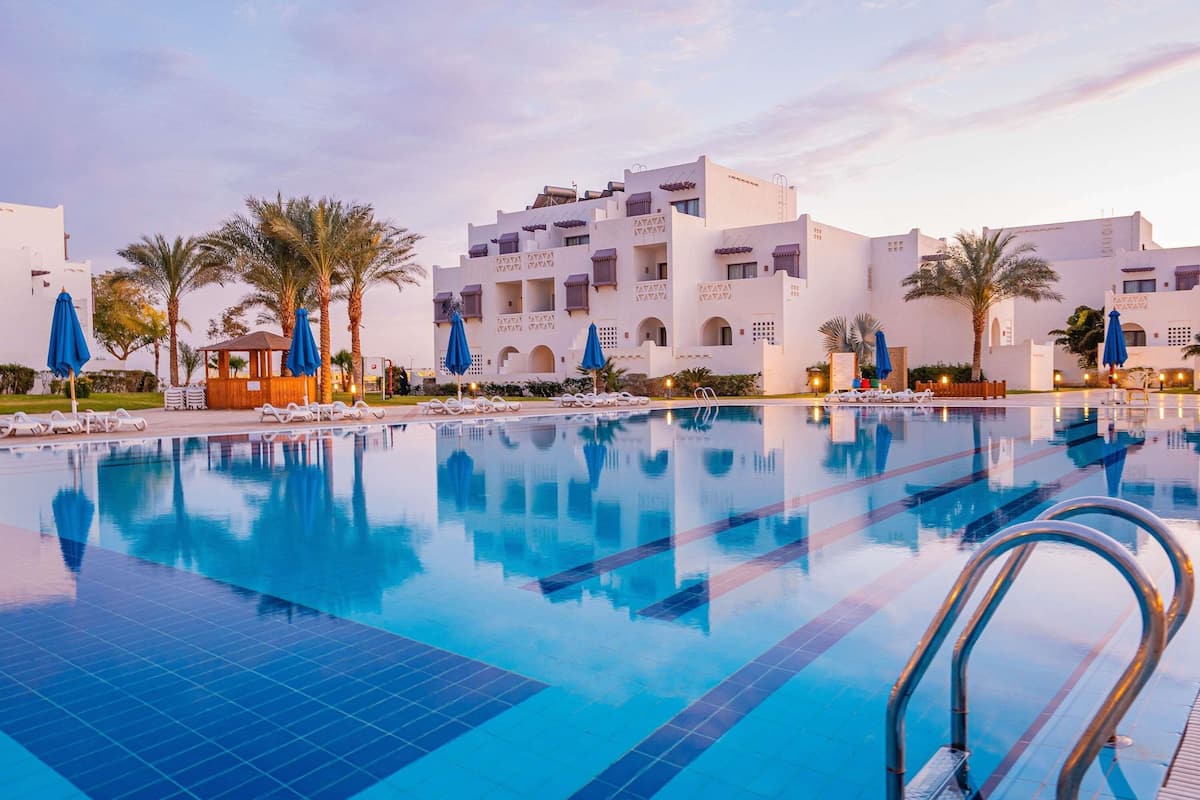Una piscina con sillas de descanso y sombrillas, rodeada de edificios blancos con balcones.