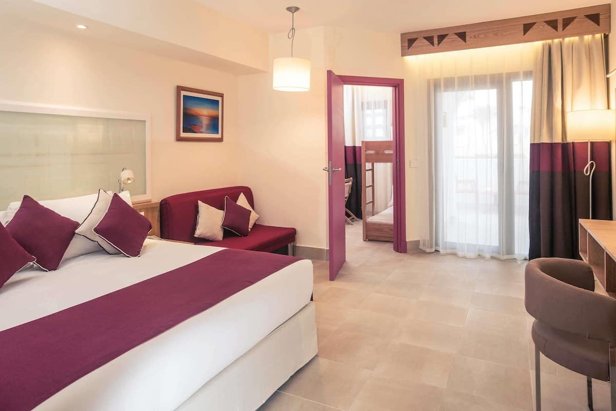 A hotel room with a bed, a desk, a chair, a window with curtains, and a door leading to another room.