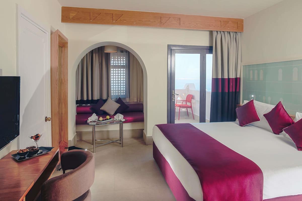 A hotel room with a bed, a TV, a desk with a tray of snacks, a sofa, and a view of the sea through a glass door.