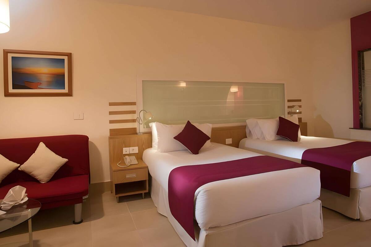 A hotel room with two beds, a red sofa, a small table, and a painting on the wall.
