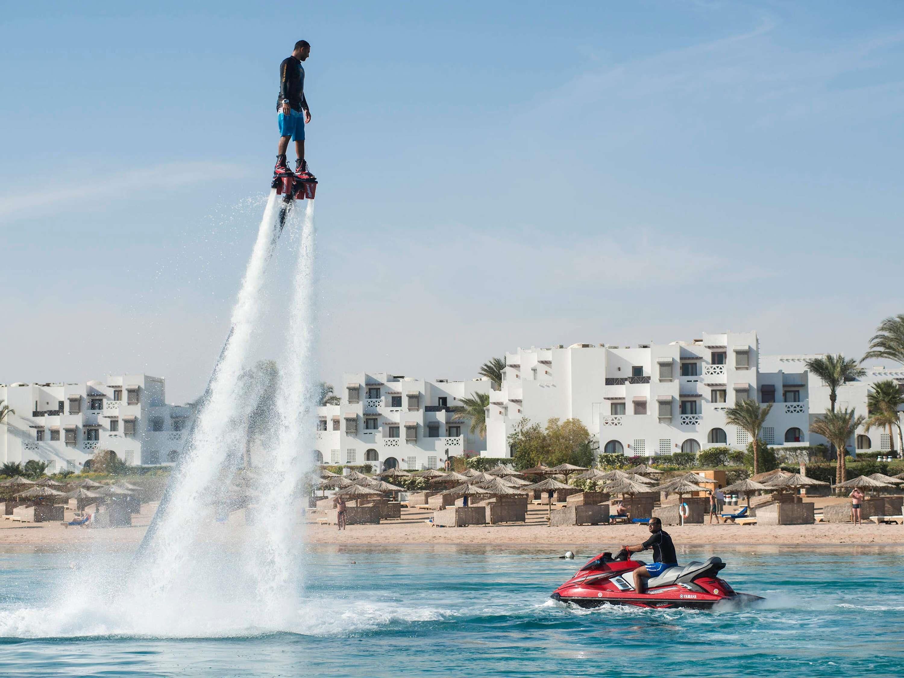Un jet ski sur l’eau, avec une personne debout sur un propulseur aquatique, et des bâtiments en arrière-plan.