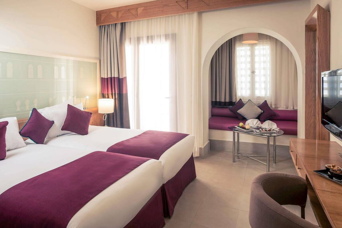 A hotel room with two beds, a TV, a small table with a lamp, and a sofa with a view.