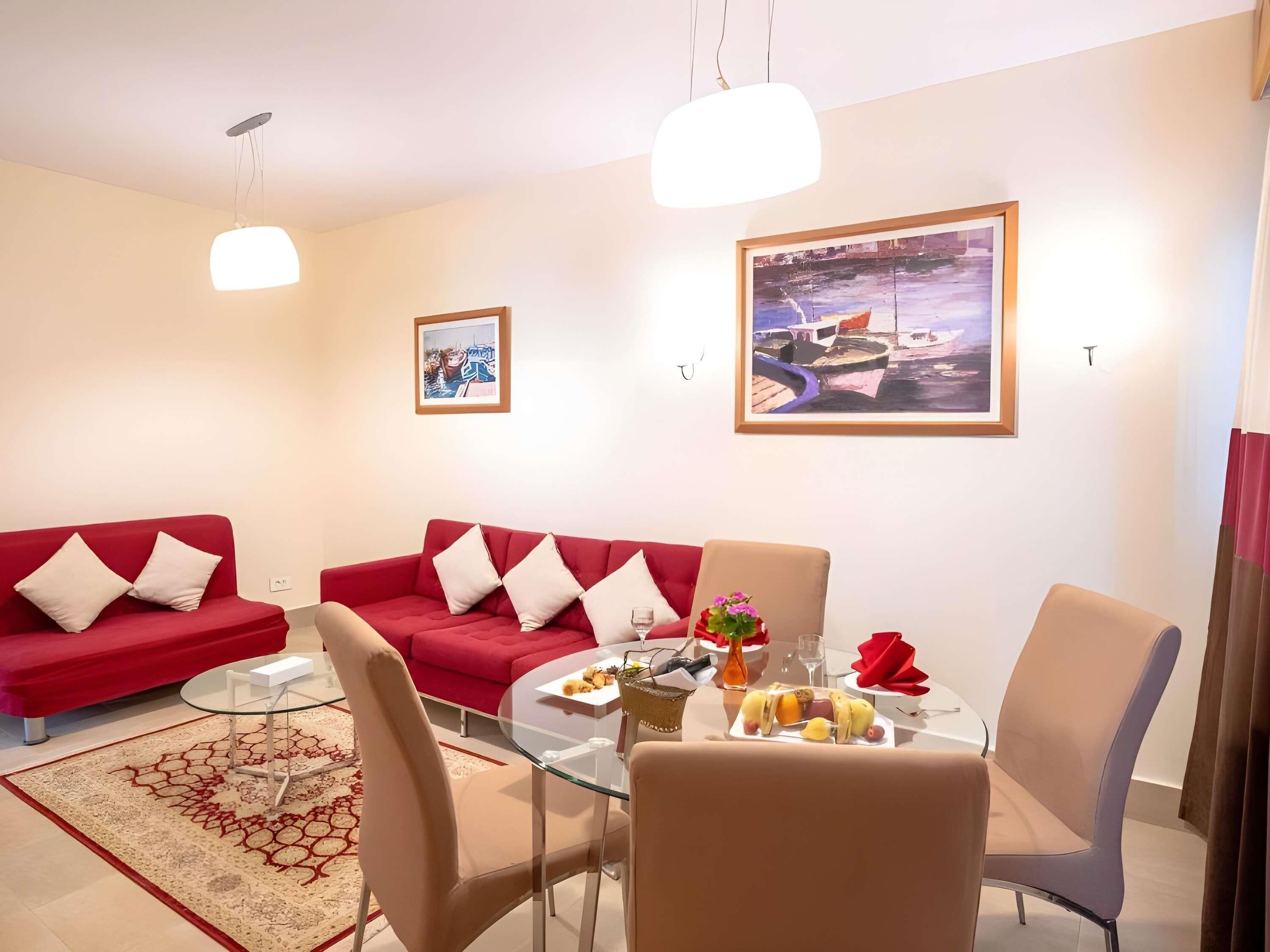 A modern living room with two red sofas, a glass dining table, and a painting of a harbor scene.