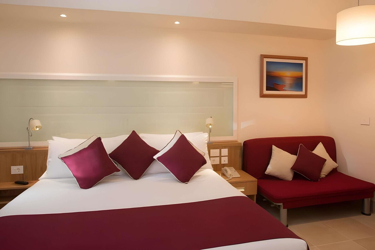 A hotel room with a large bed, a red sofa, a nightstand with a lamp, and a painting on the wall.
