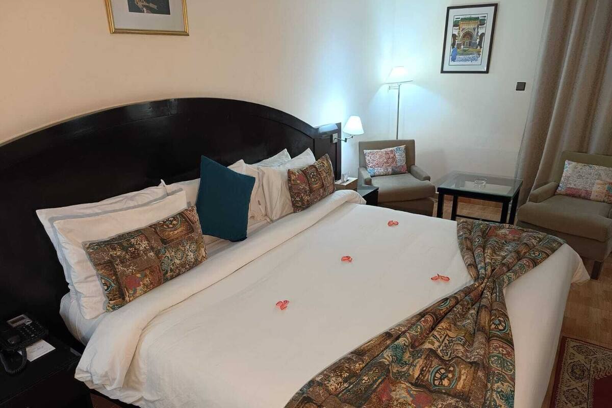 A hotel room with a large bed, a nightstand, a chair, a desk, and a lamp.