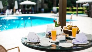 Mesa redonda con alimentos para el desayuno, una jarra y un vaso de zumo de naranja junto a una piscina.
