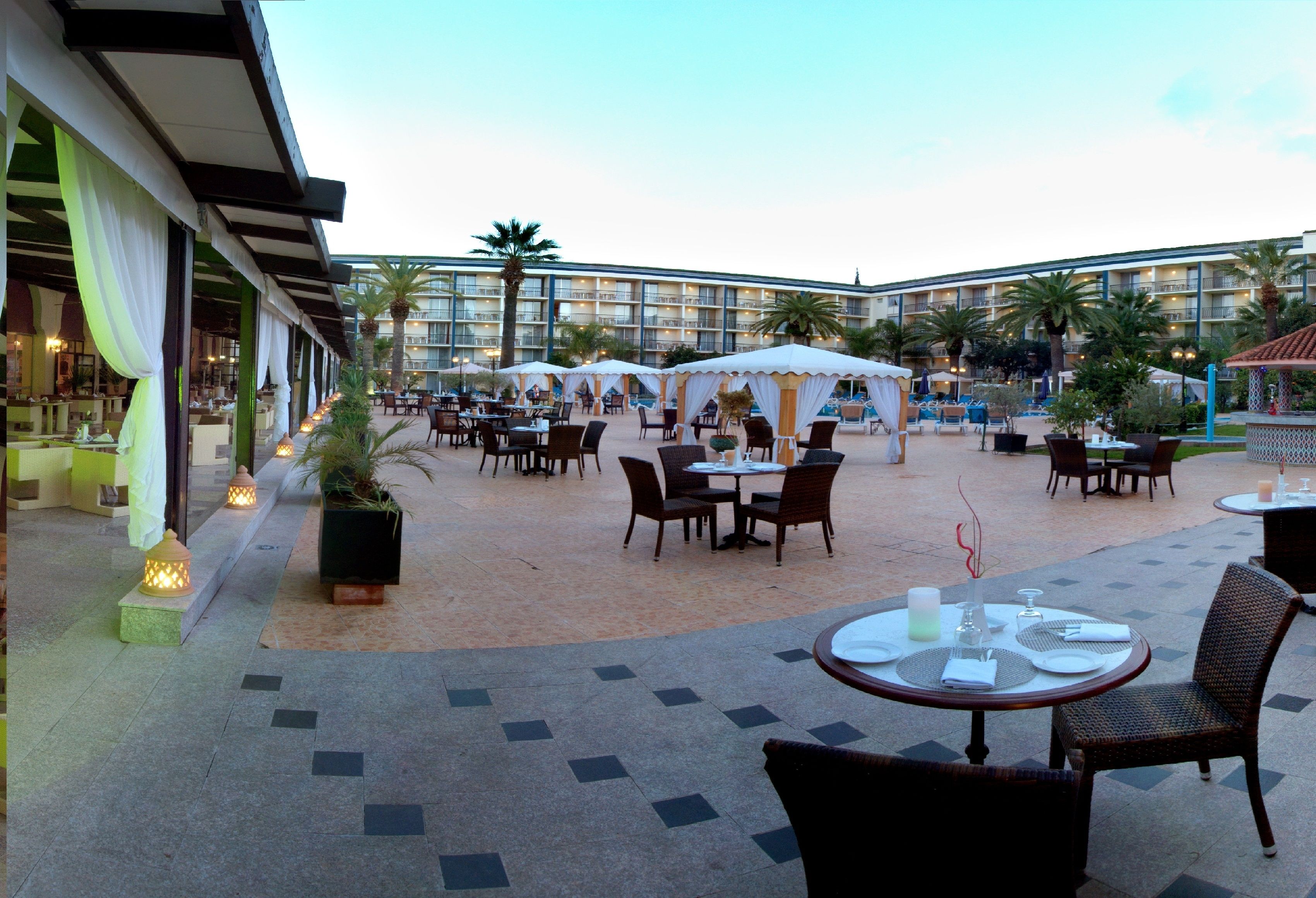 An outdoor dining area with tables and chairs, a pool in the background, and a hotel building with multiple floors.