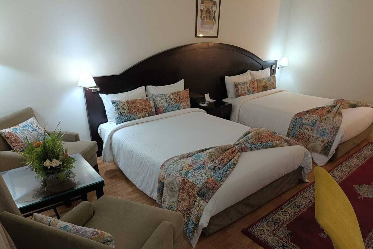 A hotel room with two beds, a sofa, a coffee table with a floral arrangement, and a red rug.