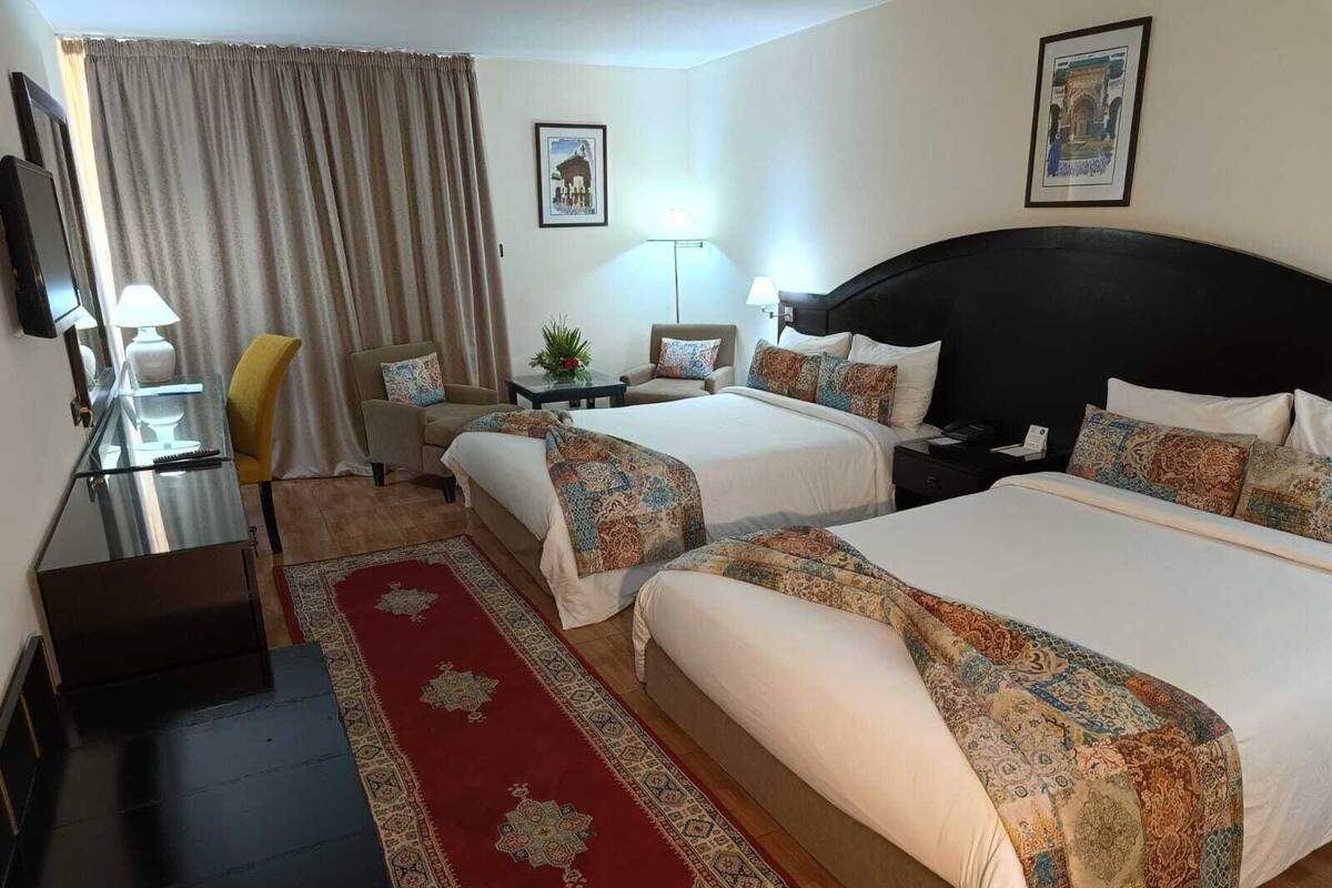 A hotel room with two beds, a desk, a chair, a TV, and a red carpet.