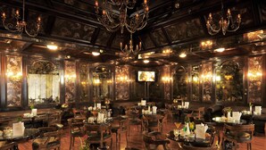 Un comedor lujoso con varias mesas y sillas, candelabros ornamentales y un espejo grande que refleja la decoración del salón.
