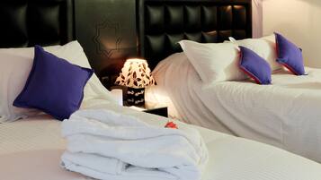 Habitación de hotel con dos camas, cada una con cabecero, sábanas blancas y almohadas moradas. Entre las camas, una mesita de noche con lámpara.