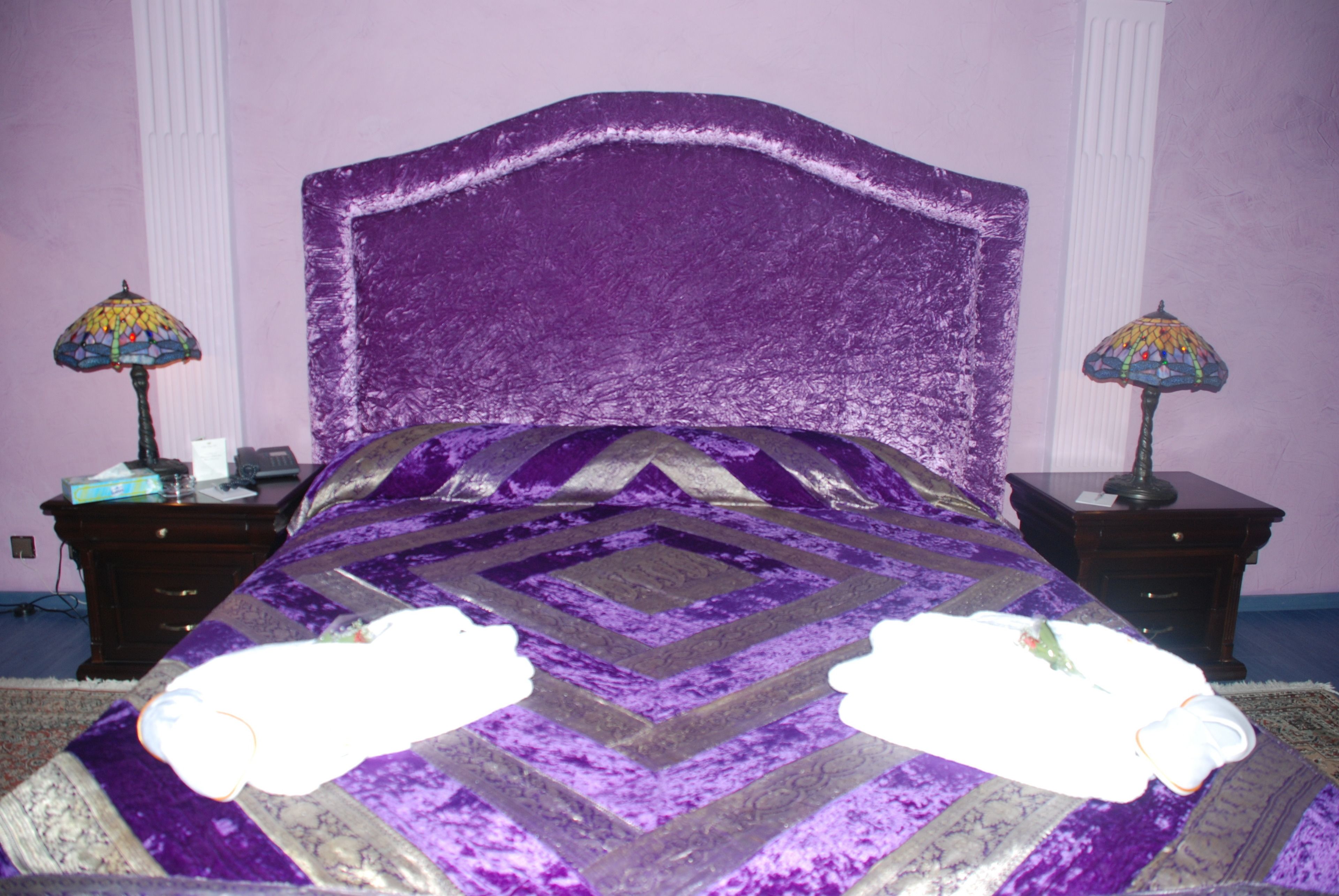 Una cama morada con una colcha estampada, dos mesitas de noche con lámparas y una alfombra blanca.