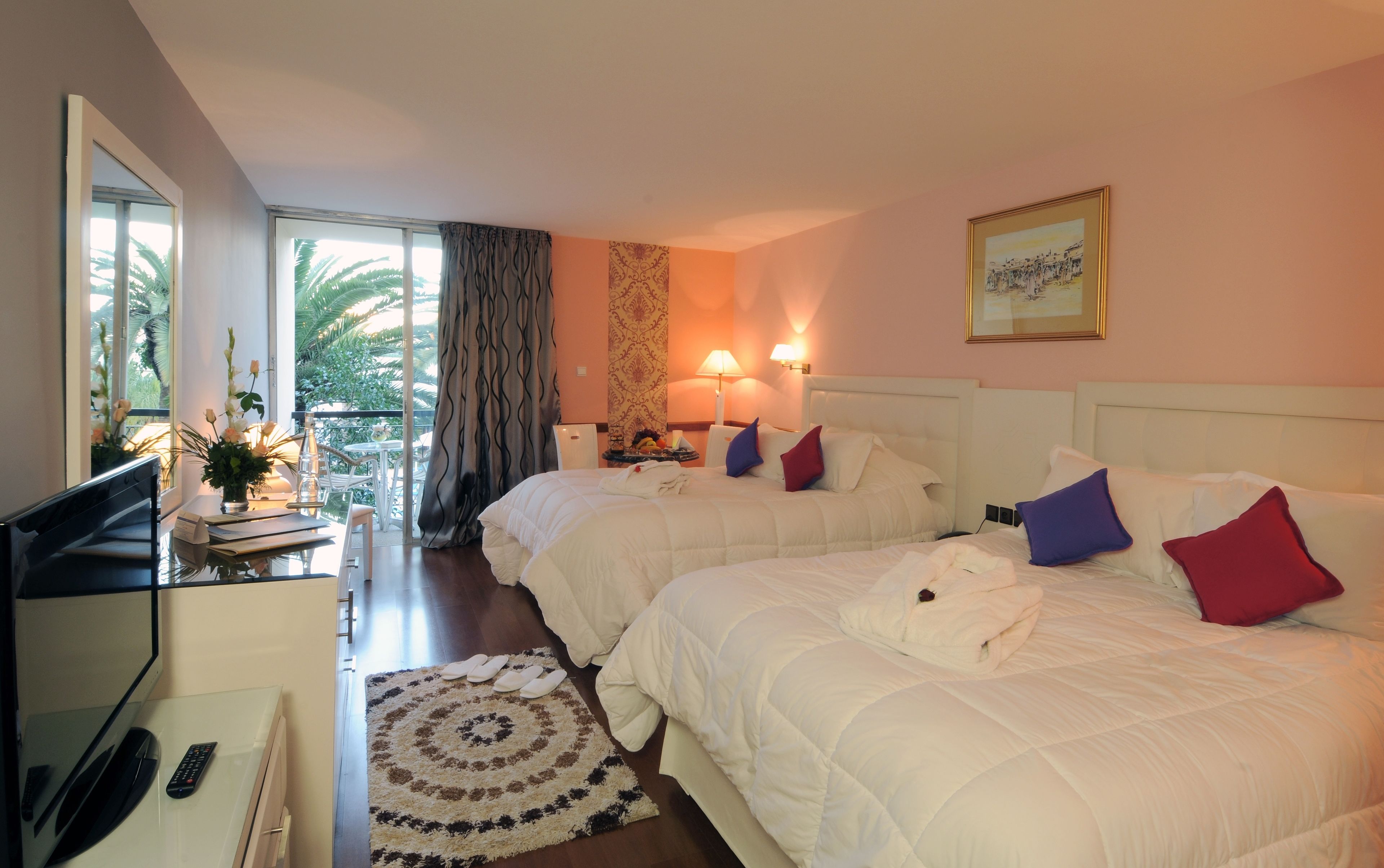 A hotel room with two beds, a desk with a TV, a balcony with a view of palm trees, and a painting on the wall.