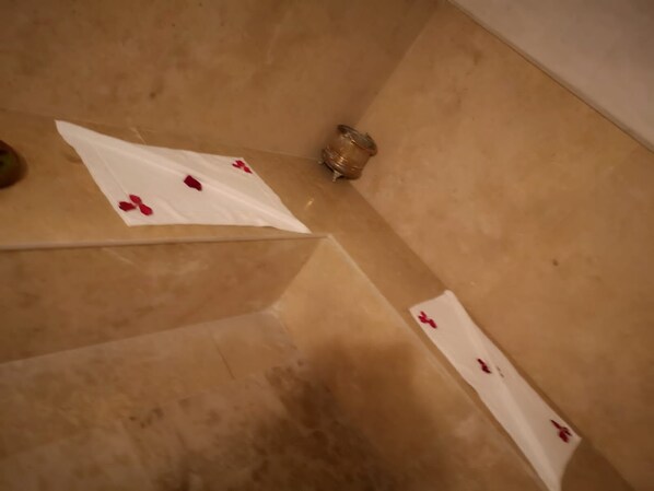 Una esquina de una habitación con una pared blanca y un suelo color beige. Se observa un objeto blanco con marcas rojas, y un pequeño recipiente en el suelo.