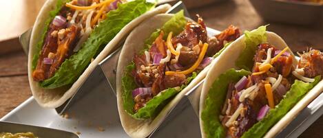 Three tacos with lettuce, red onions, and shredded cheese on a white tray, accompanied by a glass of iced beverage and lime wedges.