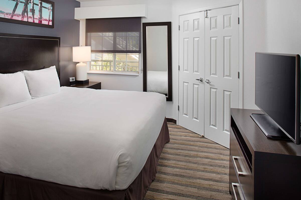 Una habitación de hotel con una cama grande, una mesita de noche con lámpara, un televisor en una cómoda y una puerta con espejo.