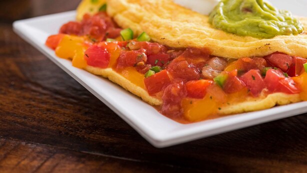 Une assiette de petit-déjeuner avec une omelette, des tomates et du guacamole, le tout servi sur une assiette blanche.