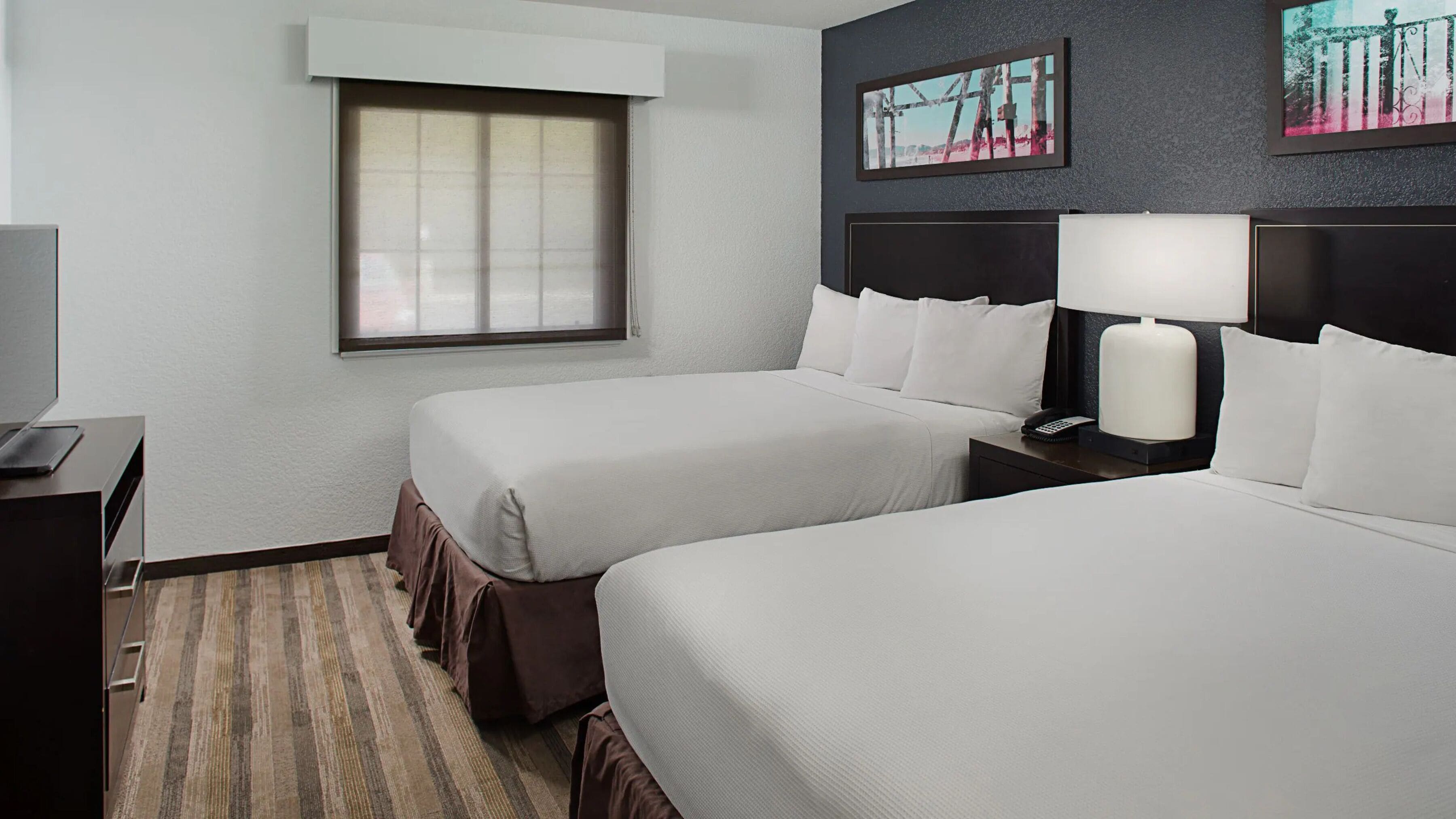 A hotel room with two beds, a desk, a lamp, and a window.