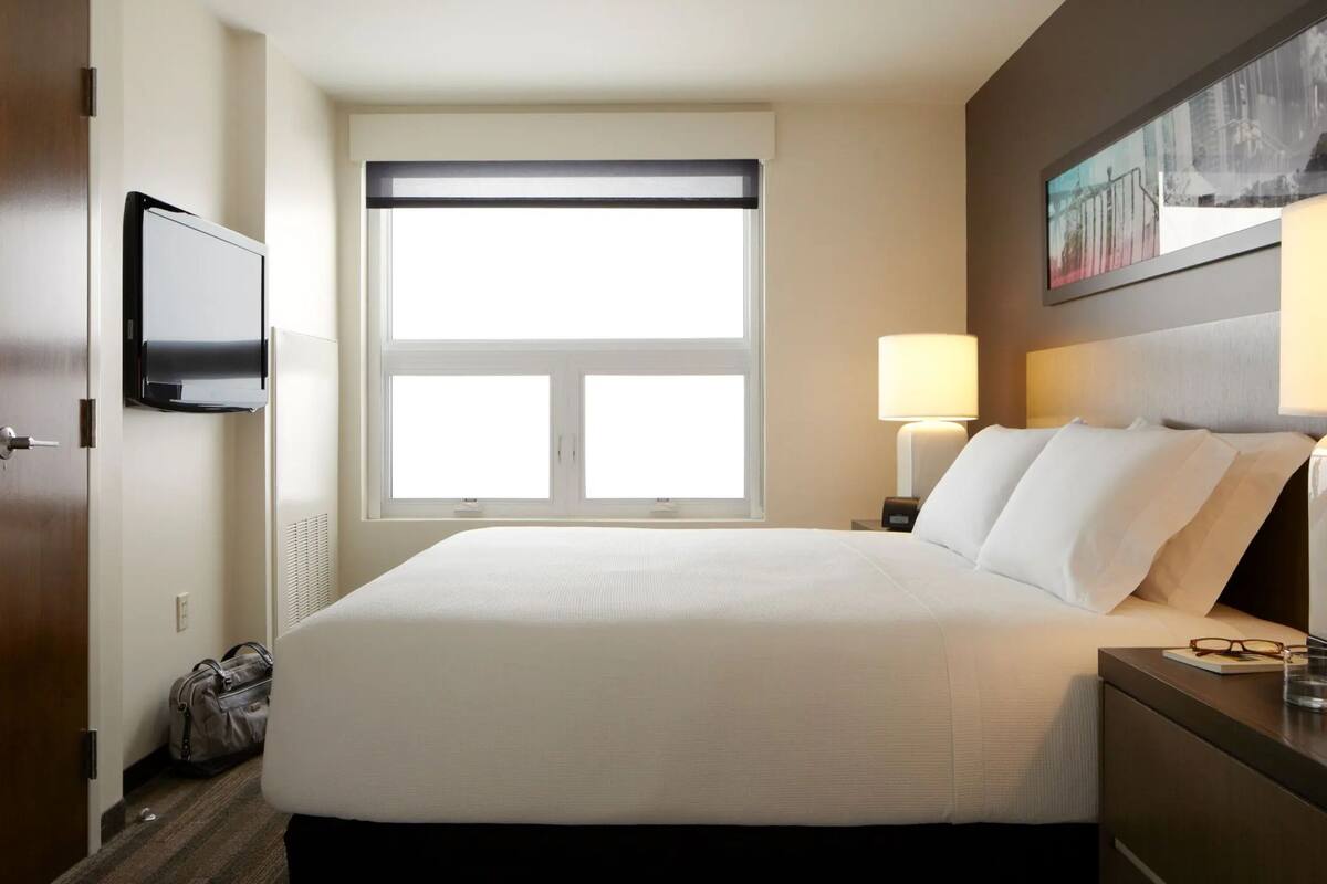 Habitación de hotel con una cama grande, dos lámparas de noche, un televisor de pantalla plana en la pared, una ventana con persianas y un cuadro enmarcado sobre la cama.