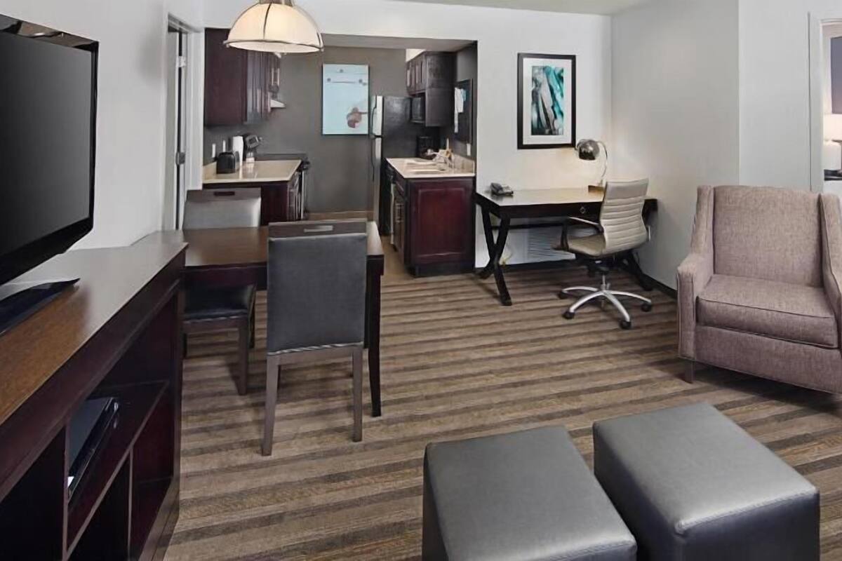 Habitación de hotel con televisión, escritorio, silla, cama y un pequeño reposapiés.
