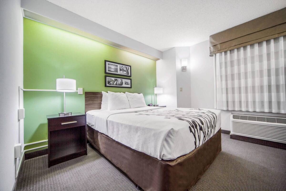 A hotel room with a bed, nightstand, wall-mounted lamps, and a window with blinds.