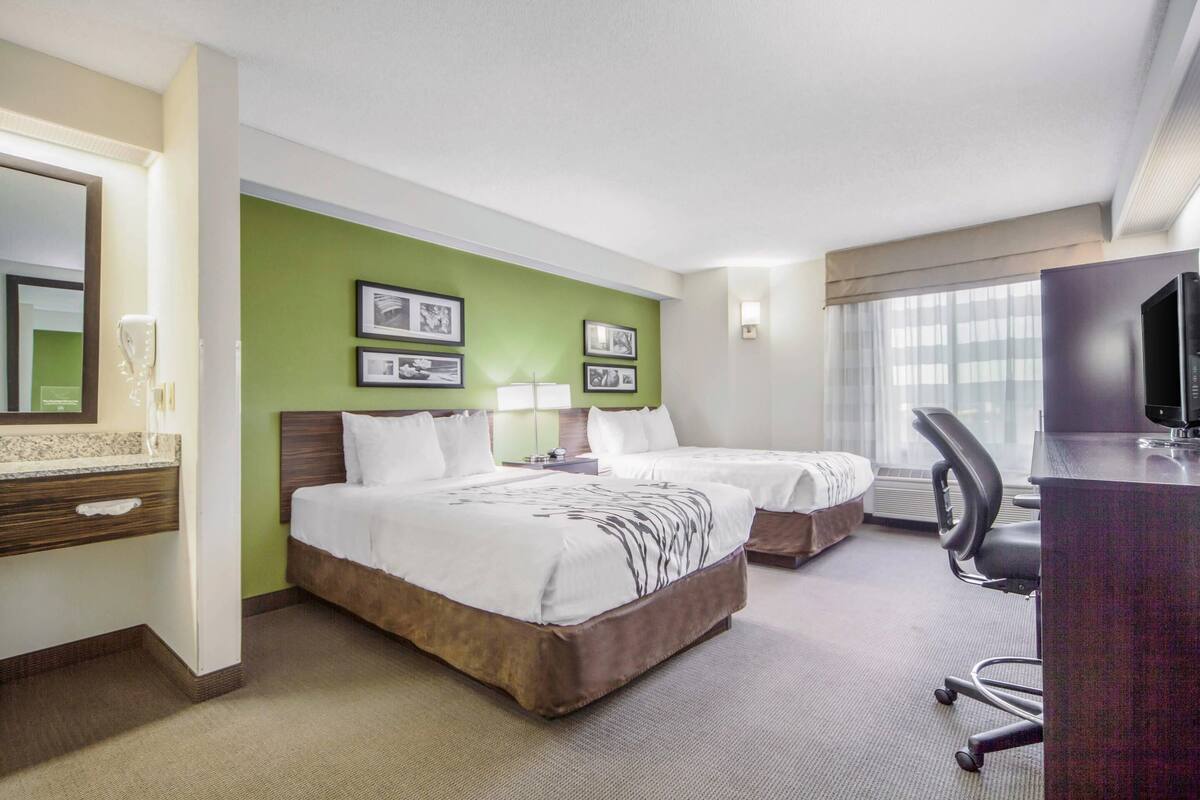 A hotel room with two beds, a desk with a chair, a TV, and framed pictures on the wall.