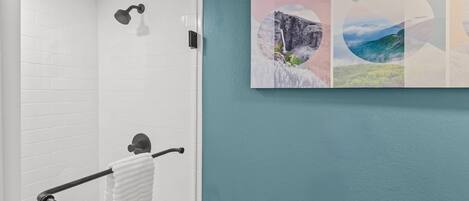 A bathroom with white tiled walls, a towel rack with a white towel, a showerhead, and a wall-mounted towel dispenser.