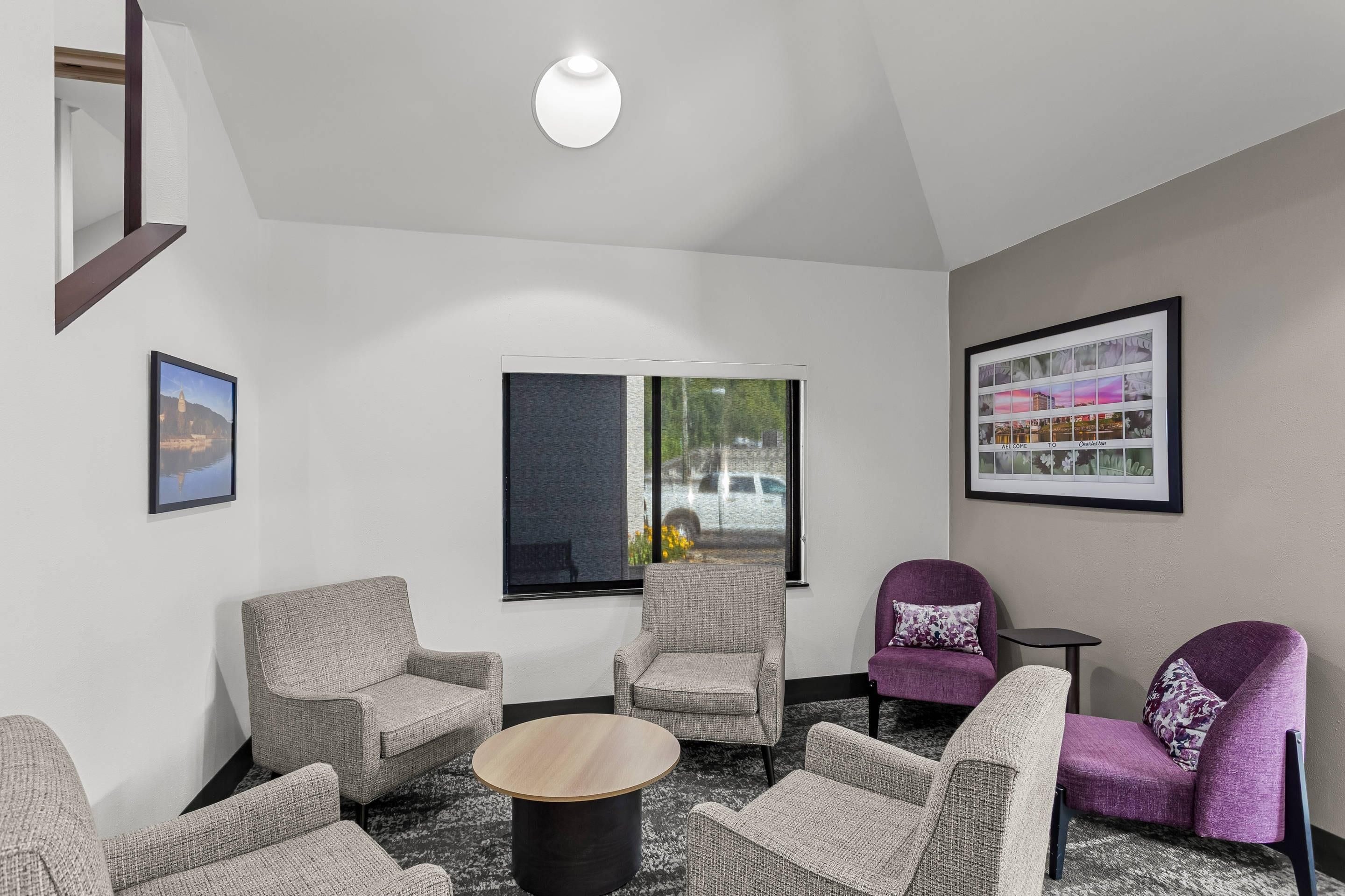 A modern lounge area with a round coffee table, grey upholstered chairs, a purple armchair, and a framed cityscape picture on the wall.