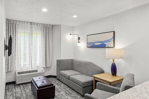 A hotel room with a grey sofa, a small wooden table, a lamp, a television, and a window with curtains.