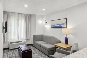 Quarto de hotel com sofá cinzento, uma pequena mesa de madeira, luminária, televisão e janela com cortinas.