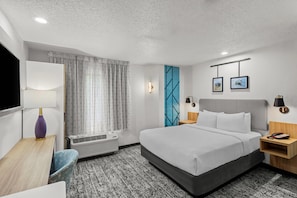 Quarto de hotel com cama, mesas de cabeceira, escrivaninha, cadeira, televisão na parede, janela com cortinas e tapete estampado.