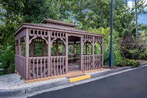 Um gazebo de madeira com varanda coberta, rodeado por vegetação e um caminho pavimentado.