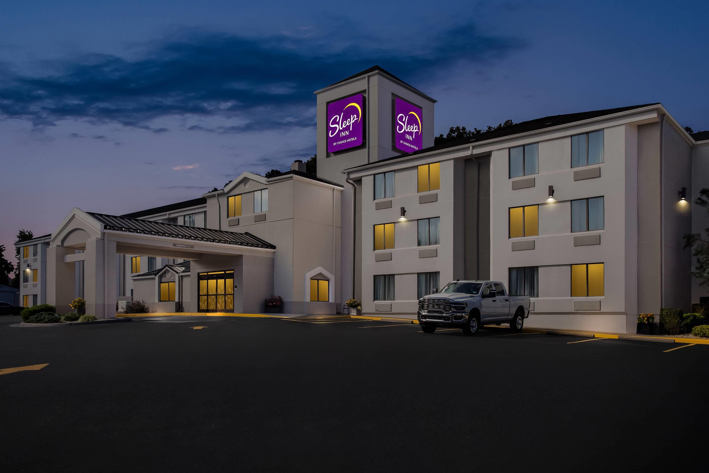 Un hôtel Sleep Inn au design moderne, doté d’un grand parking et d’un camionnette blanche garée devant l’entrée.