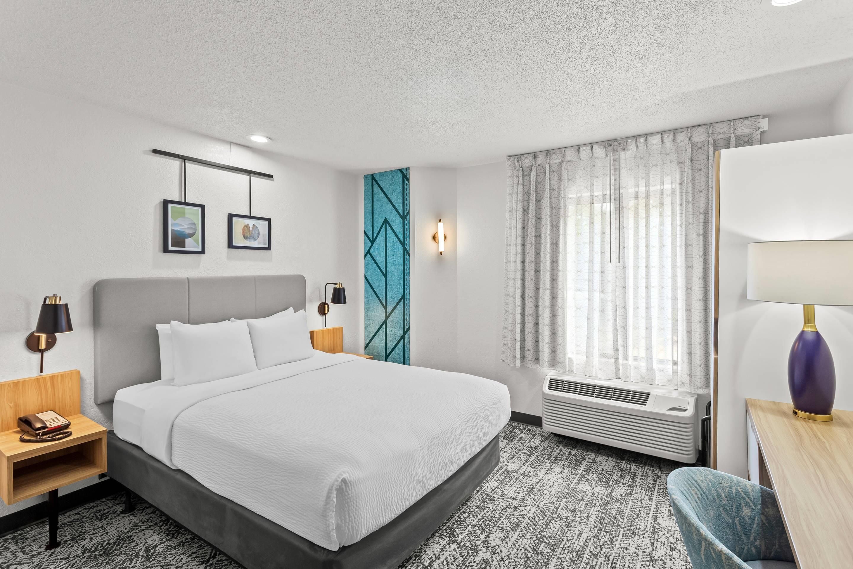 A hotel room with a bed, bedside tables, a desk with a lamp, a chair, a window with curtains, and a wall with framed pictures.
