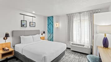 A hotel room with a bed, bedside tables, a desk with a lamp, a chair, a window with curtains, and a wall with framed pictures.
