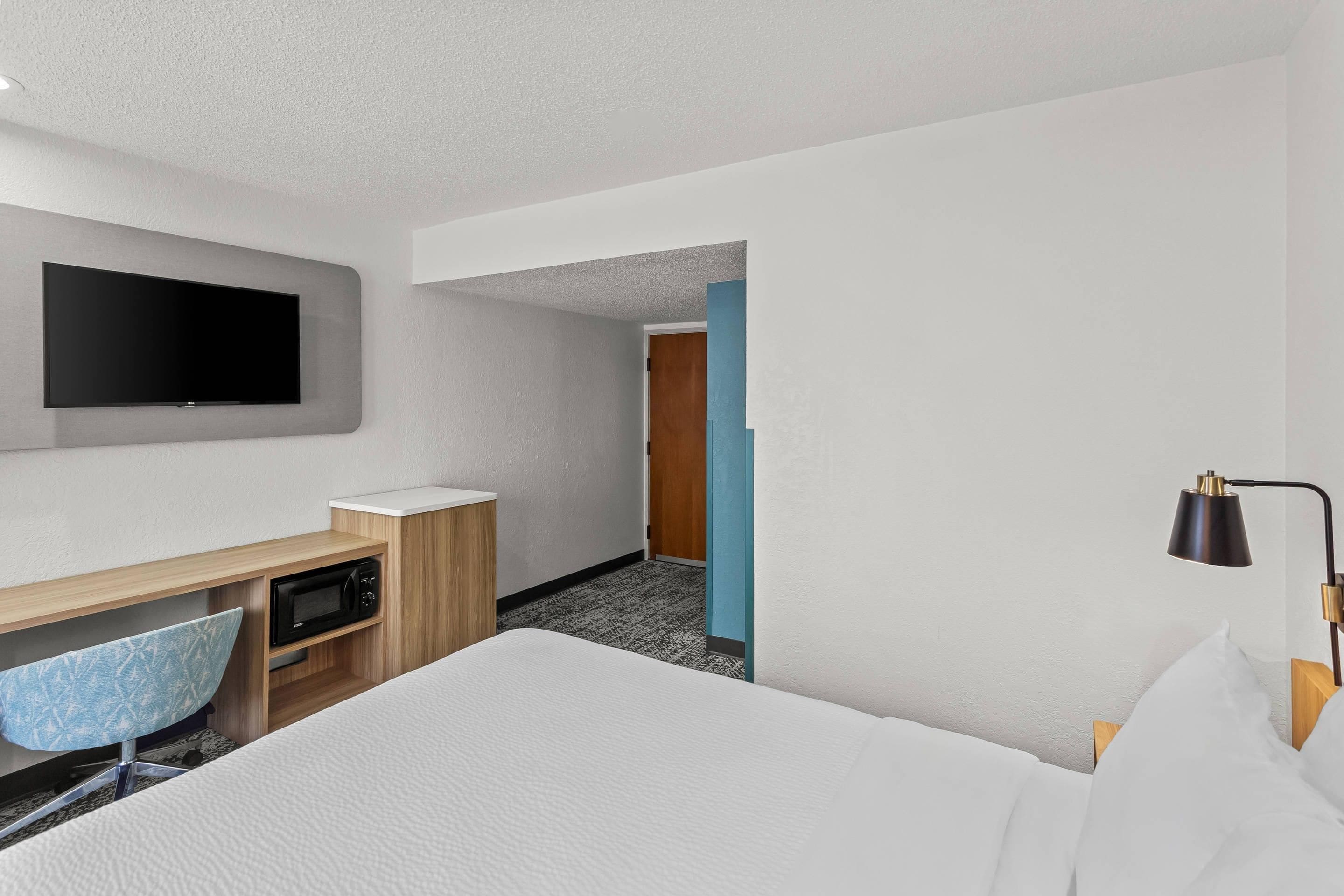 A hotel room with a bed, a desk with a flat-screen TV, a microwave, a chair, and a nightstand.