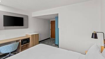 A hotel room with a bed, a desk with a flat-screen TV, a microwave, a chair, and a nightstand.