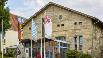 A historic stone building with a modern glass entrance, flags, and a H4 Hotel sign.