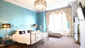 A bedroom with a four-poster bed, a chandelier, a bedside table with a lamp, a bench, and a large window with curtains. - The Victoria Hotel Manchester by Compass Hospitality (Oldham)