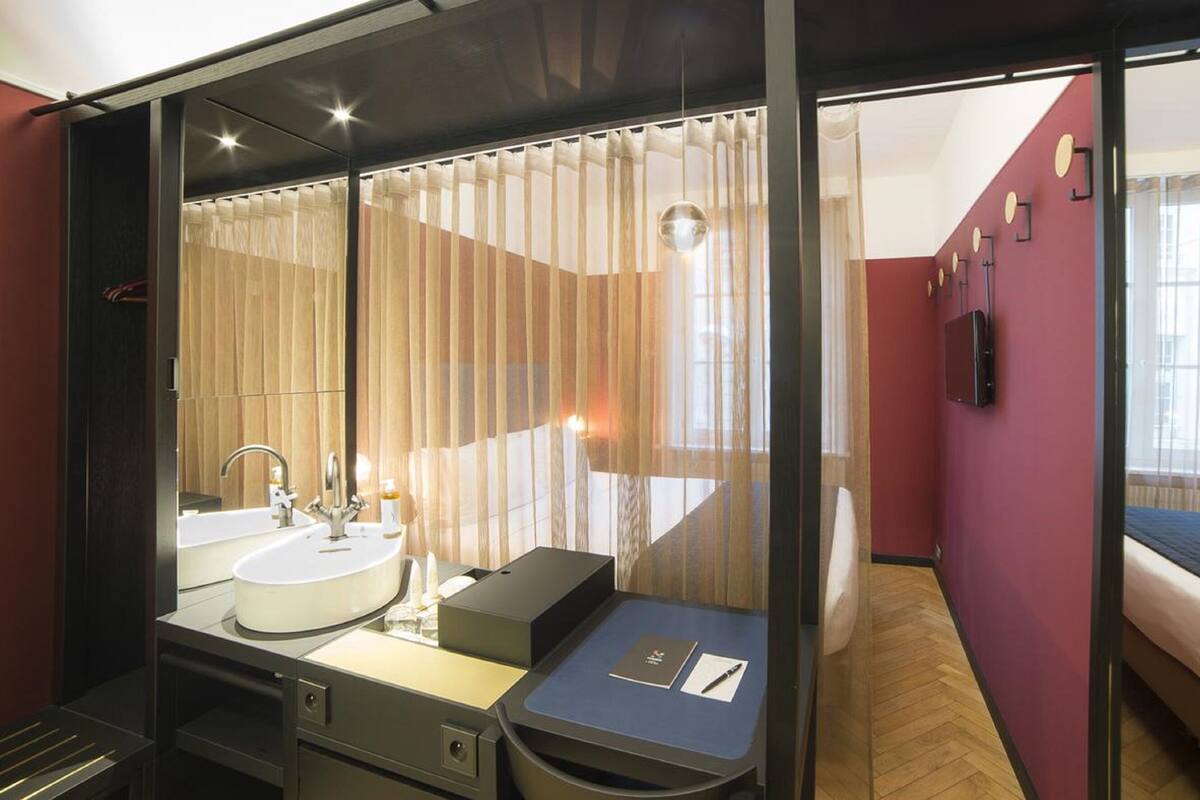 A modern hotel room with a large mirror, dual sinks, a small desk, and a bed.