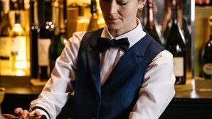 A person in a bar setting, wearing a formal vest and bow tie, with a bar counter and various bottles in the background. - Hotel Hannong (Strasbourg)
