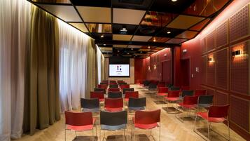 Une salle de conférence avec des rangées de chaises, un écran de projection et un mur rouge orné de panneaux décoratifs.