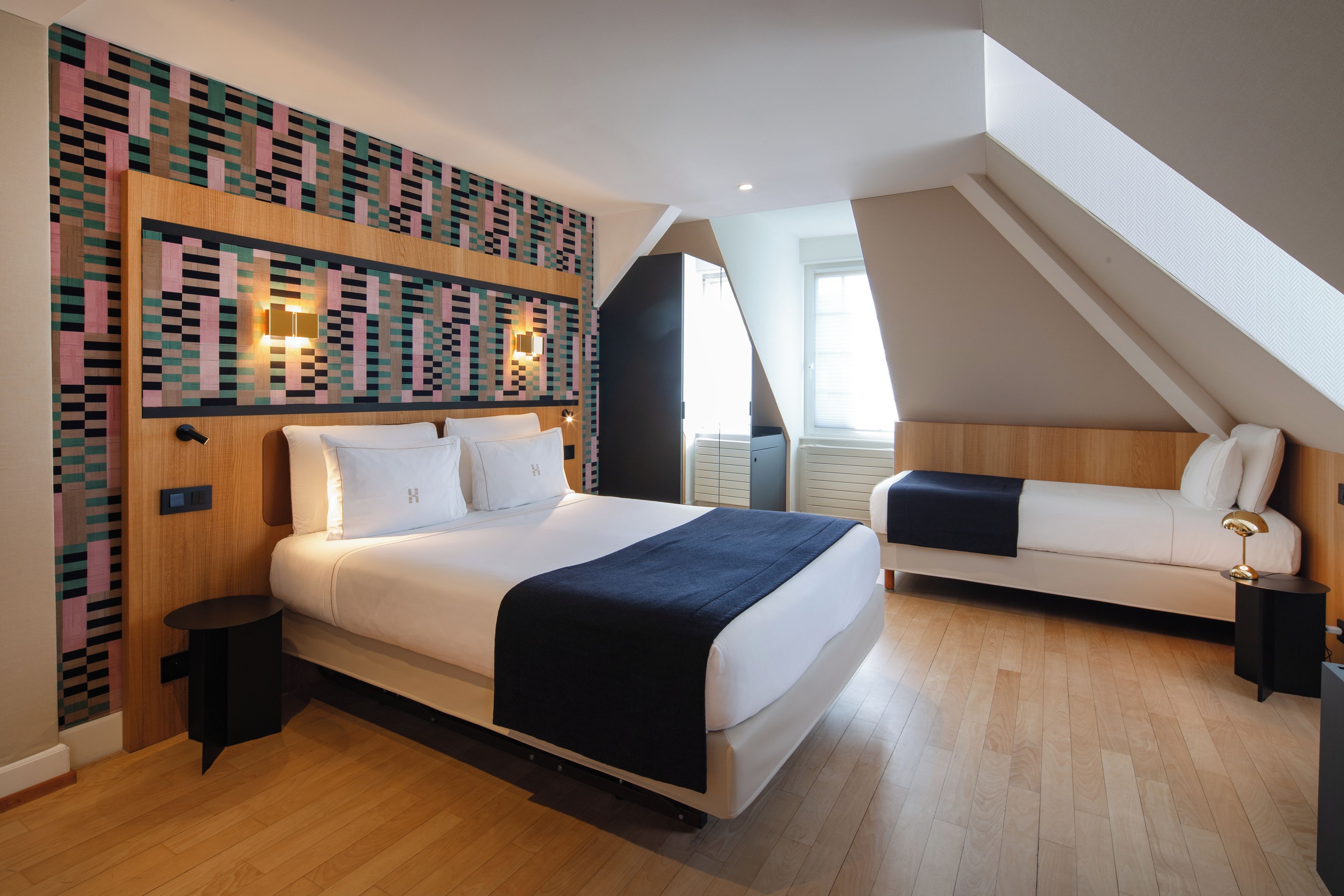 A modern bedroom with a large bed, a smaller sofa, a patterned wall, and a skylight.