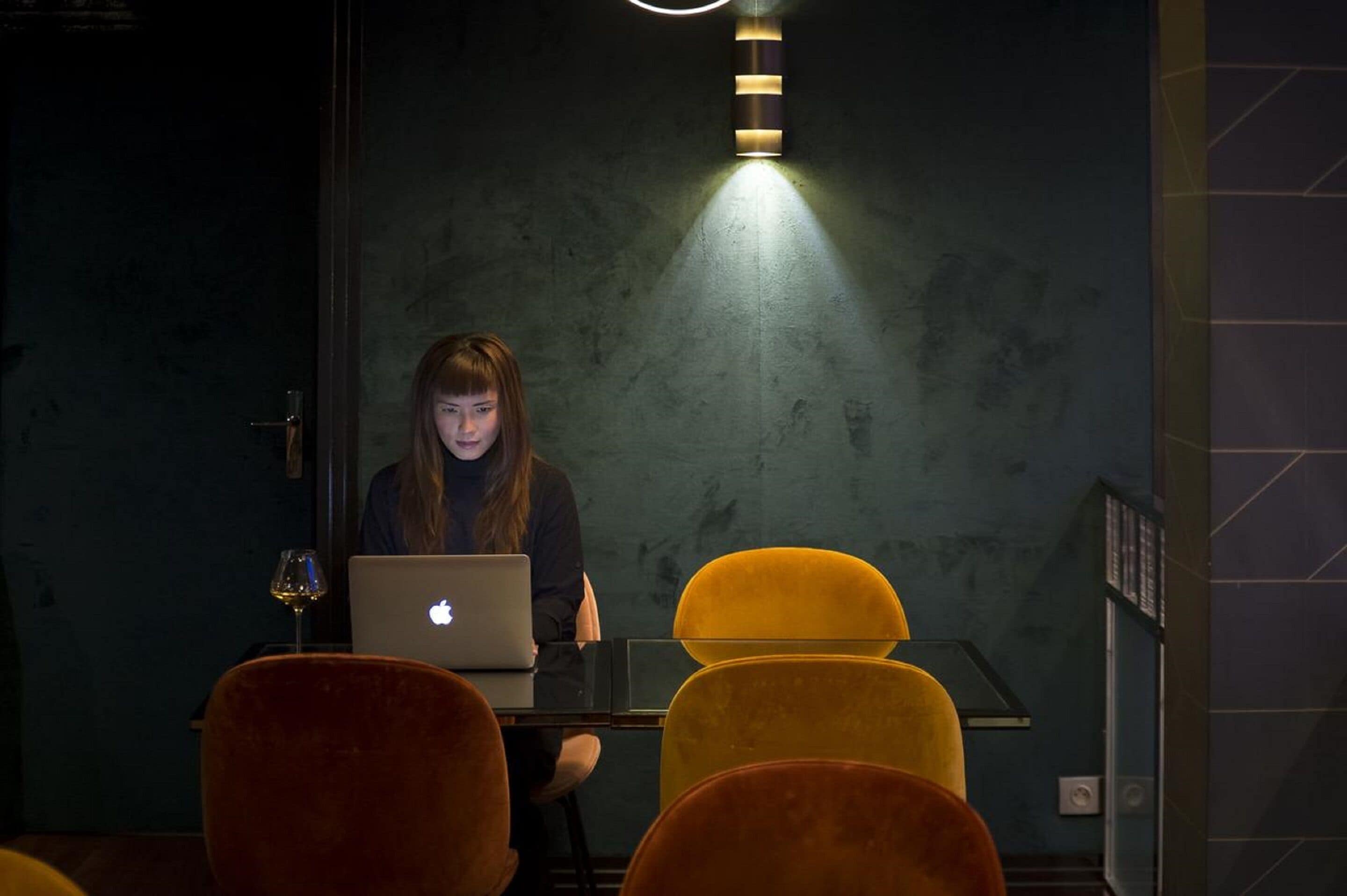 A person working on a laptop in a dimly lit room with a single light source above.