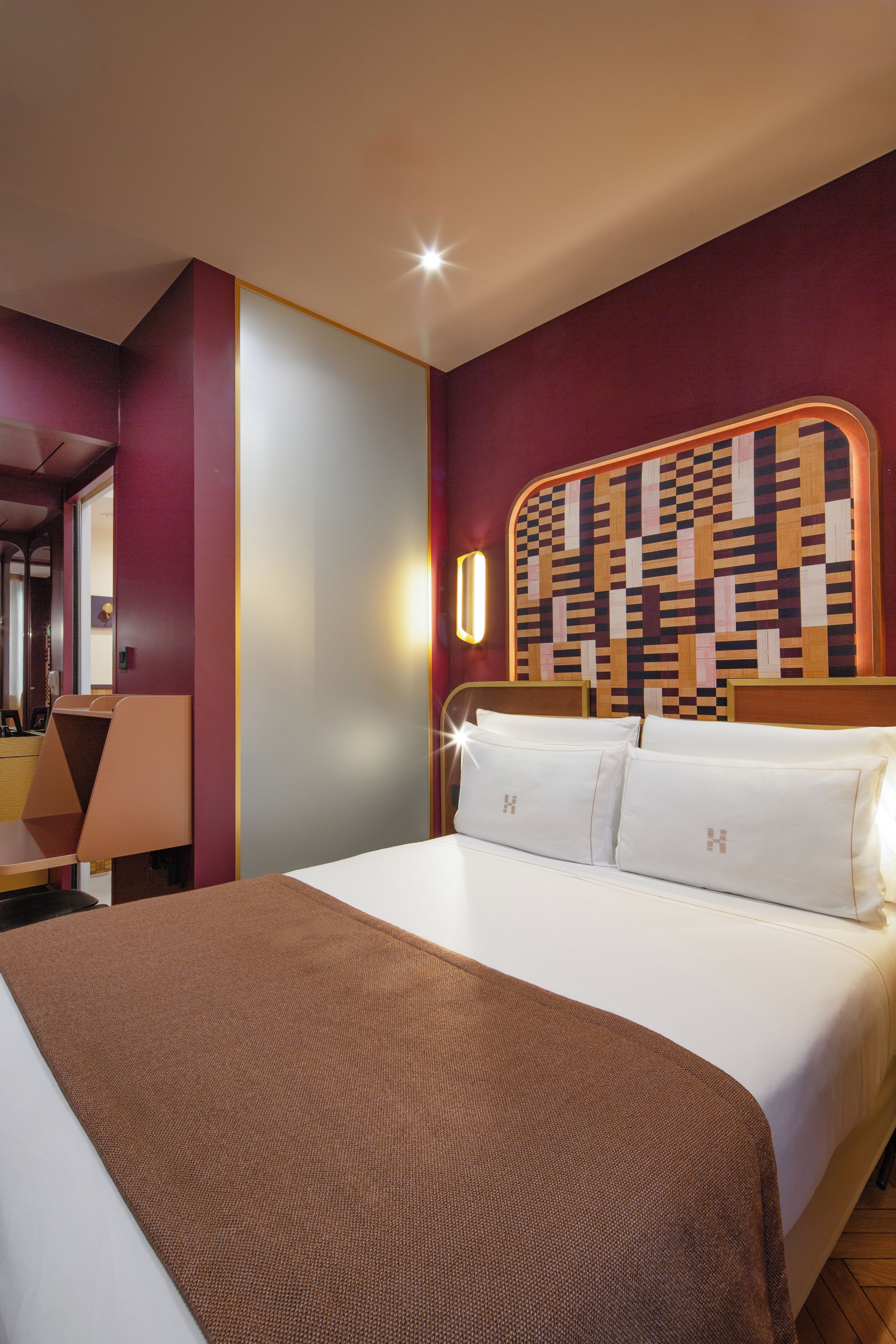 Une chambre d’hôtel avec un grand lit, une tête de lit ornée d’un motif géométrique, une table de chevet avec une lampe et un miroir au mur.
