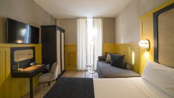 A hotel room with a bed, a desk with a chair, a television, a radiator, and a window with curtains.