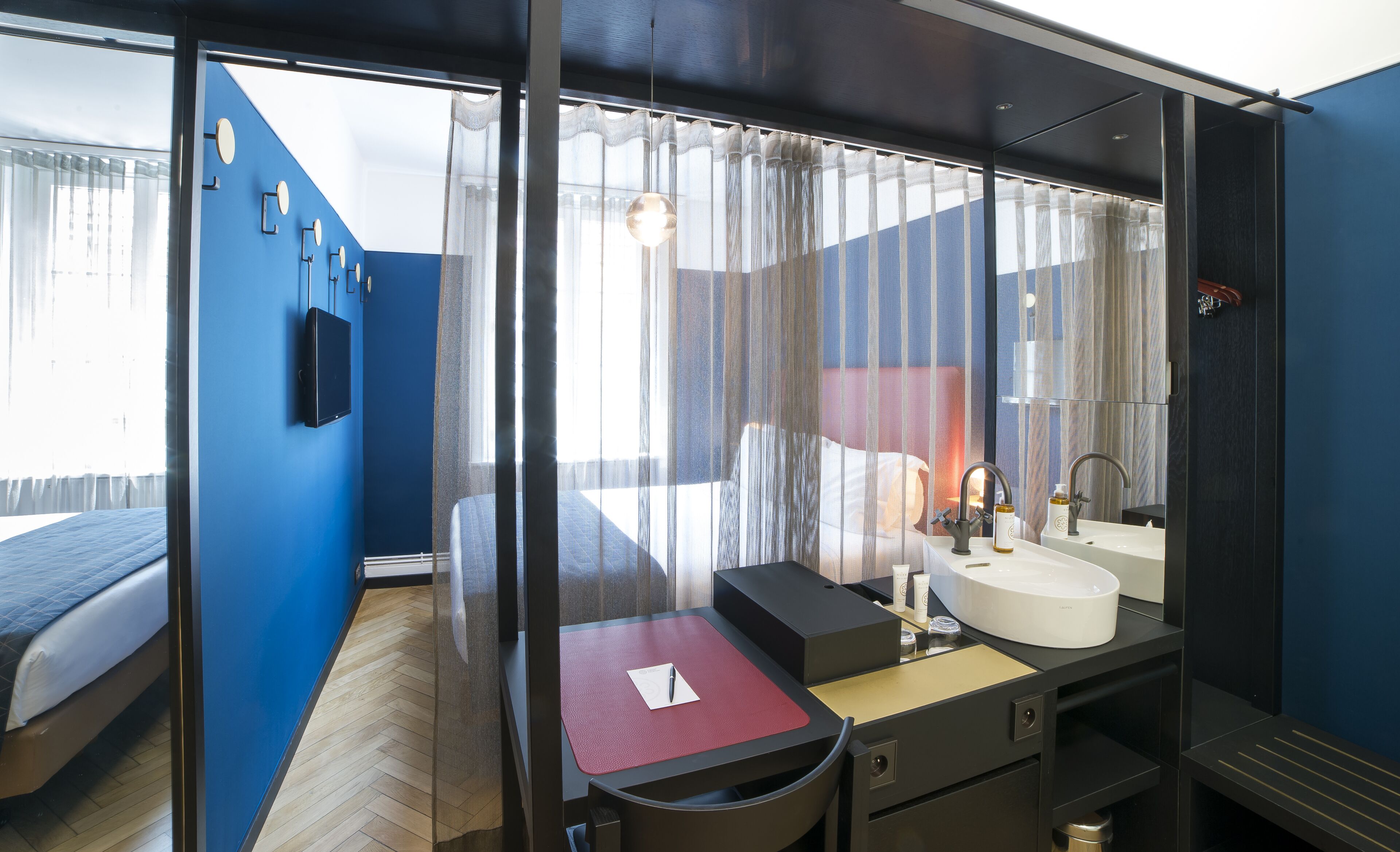 A modern hotel room with a large bed, a desk with a chair, a bathroom with a sink and mirror, and a view through sheer curtains.