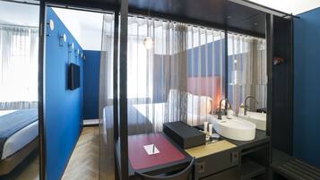 A modern hotel room with a large bed, a desk with a chair, a bathroom with a sink and mirror, and a view through sheer curtains.