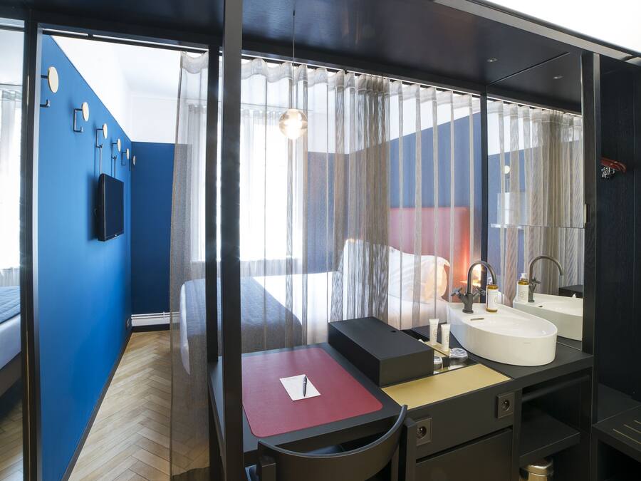 A modern hotel room with a large bed, a desk with a chair, a bathroom with a sink and mirror, and a view through sheer curtains.