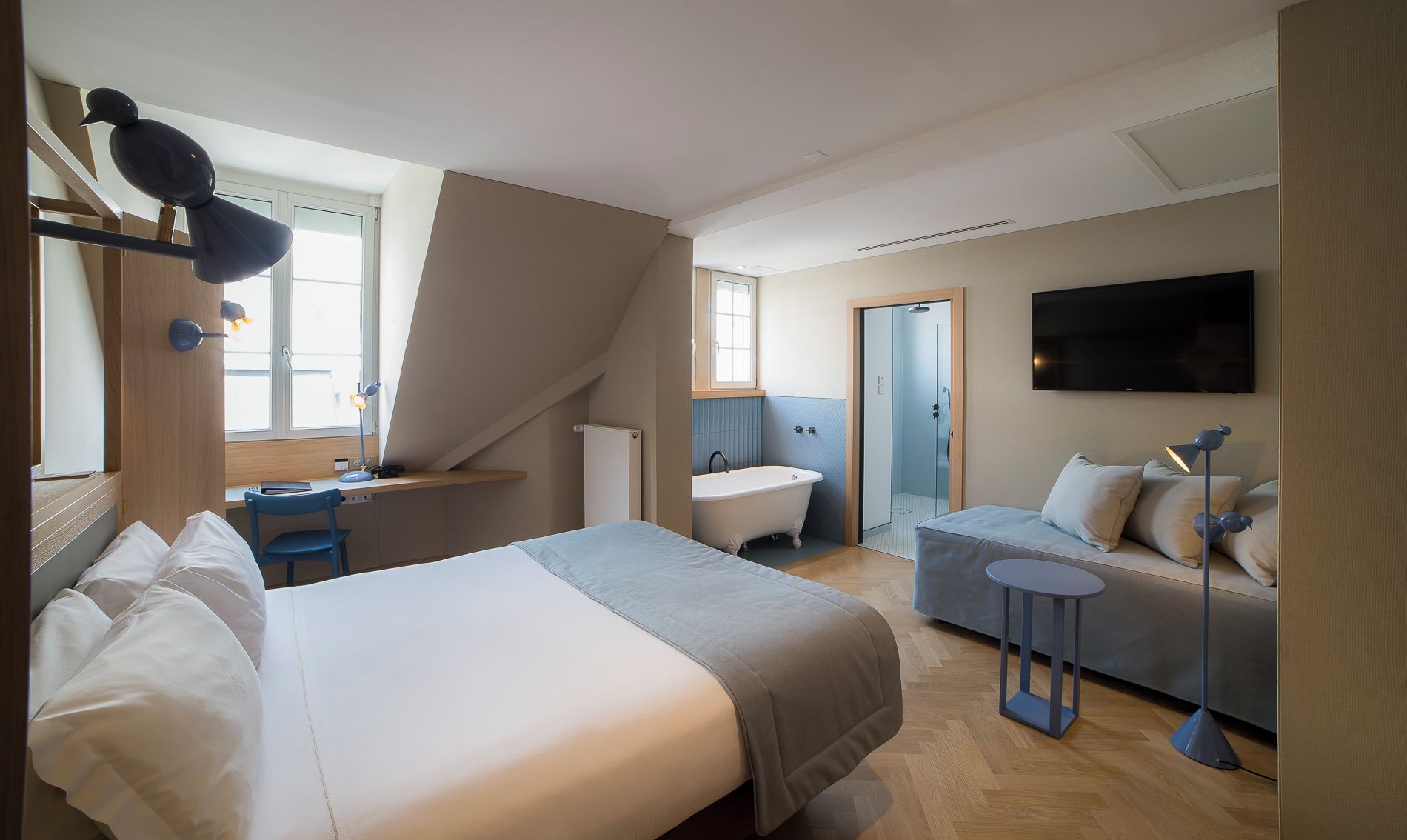 A modern hotel room with a large bed, a desk, a sofa, a TV, and a shower area.